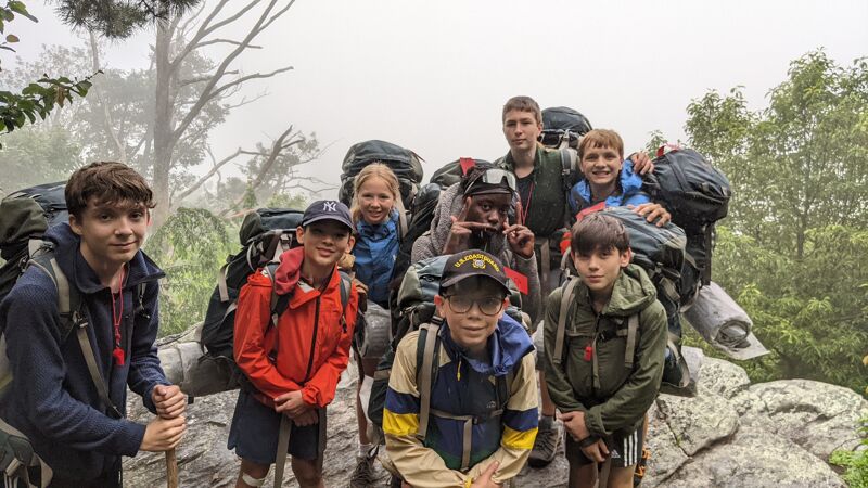 A group of young people, possibly scouts or hikers, are posing for a photo outdoors. They are wearing backpacks and outdoor clothing, suggesting they are on an adventure or expedition. The background is a bit foggy, with trees visible, indicating a natural setting, possibly a mountain or forest. The group appears to be in good spirits, smiling and looking at the camera.